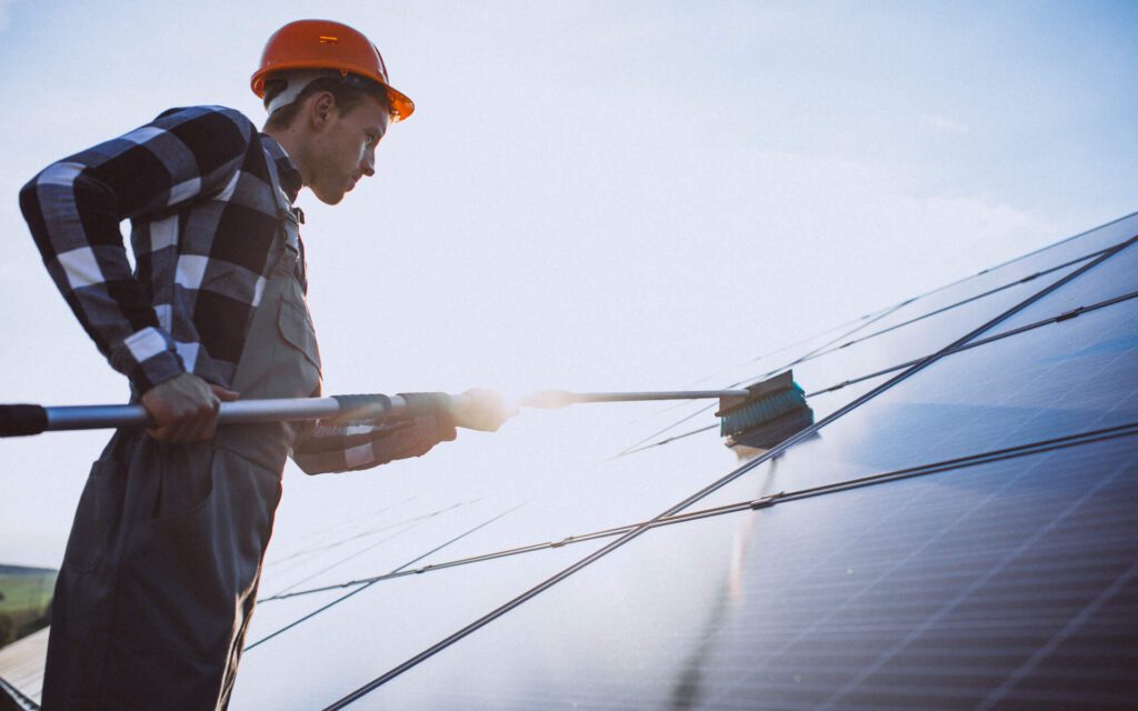 A worker wearing a hard hat performs solar panel cleaning in Los Angeles using a long-handled brush.