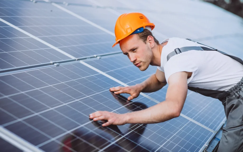 A technician wearing a hard hat performs solar panel maintenance by inspecting the surface of a solar panel array.